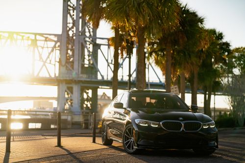 Free Sleek black luxury car parked beside palms with a bridge in the background at sunrise. Stock Photo