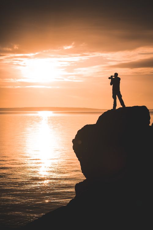 Free A stunning silhouette of a photographer taking a sunset shot on a rocky cliff by the sea, showcasing natural beauty and adventure. Stock Photo