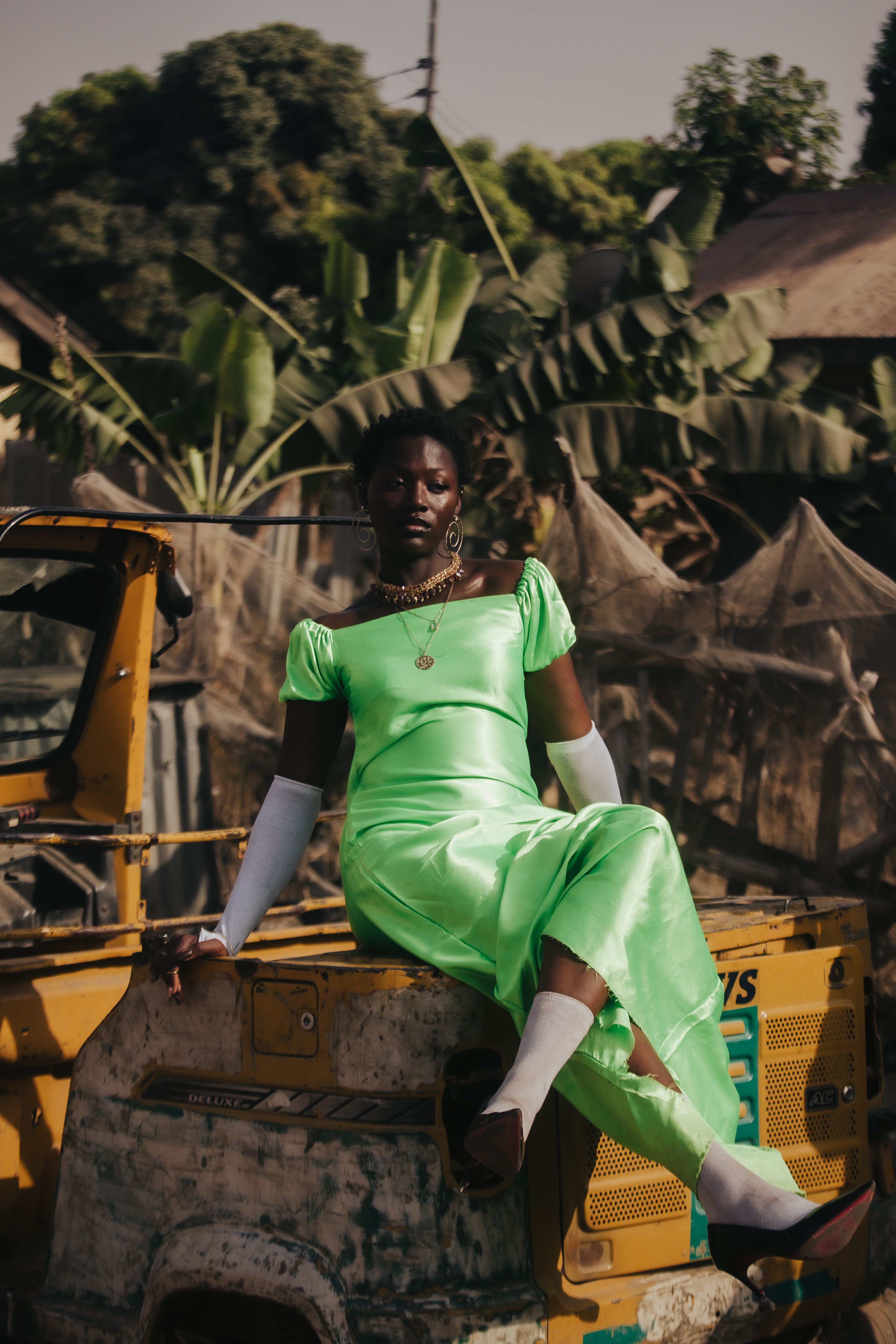 Stylish woman in vibrant green dress poses outdoors on a rustic vehicle for a fashion shoot.