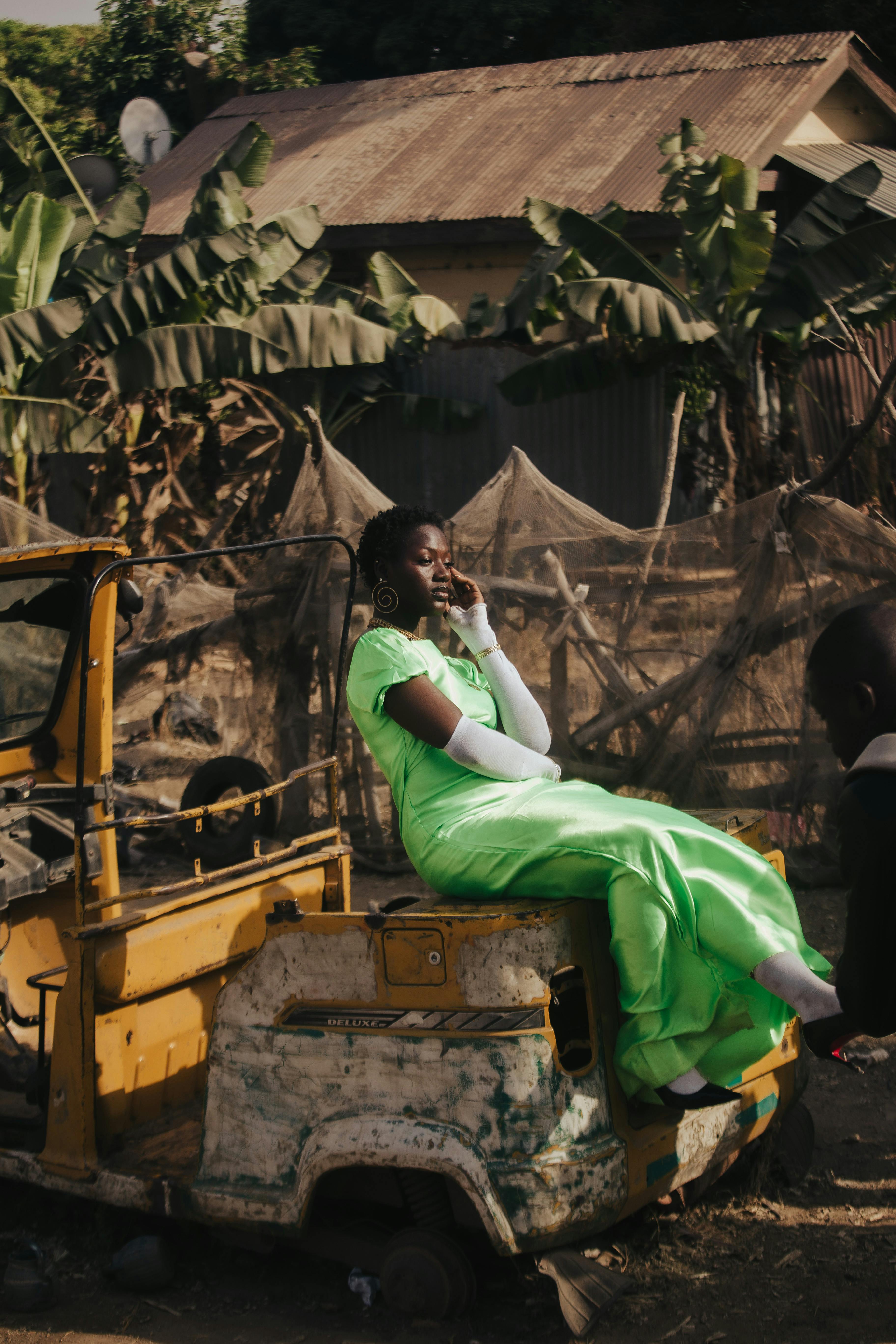 A woman in a vibrant green dress sits thoughtfully on an old vehicle, surrounded by lush greenery.