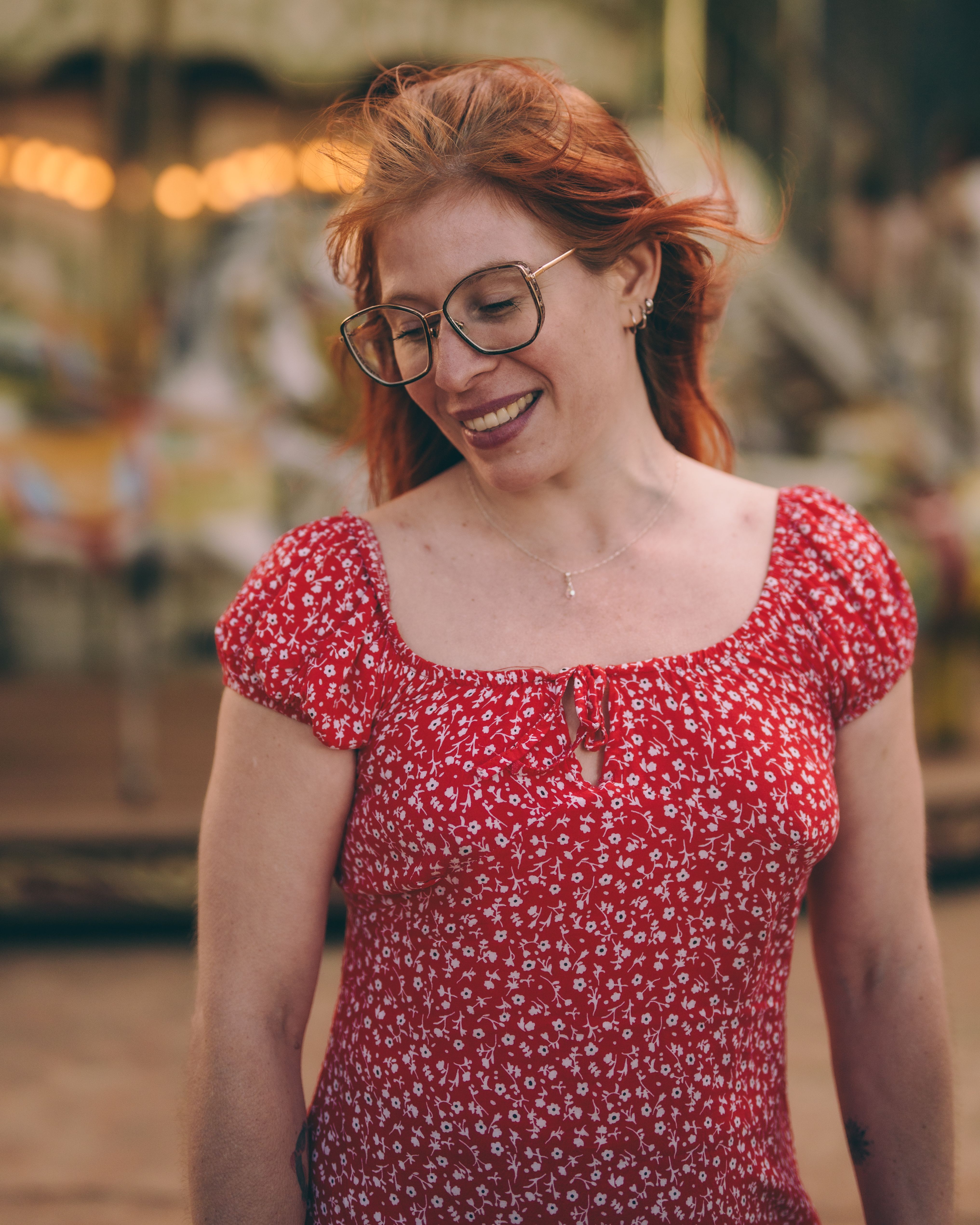 Smiling woman with red hair and glasses in a vibrant red dress at a carnival.