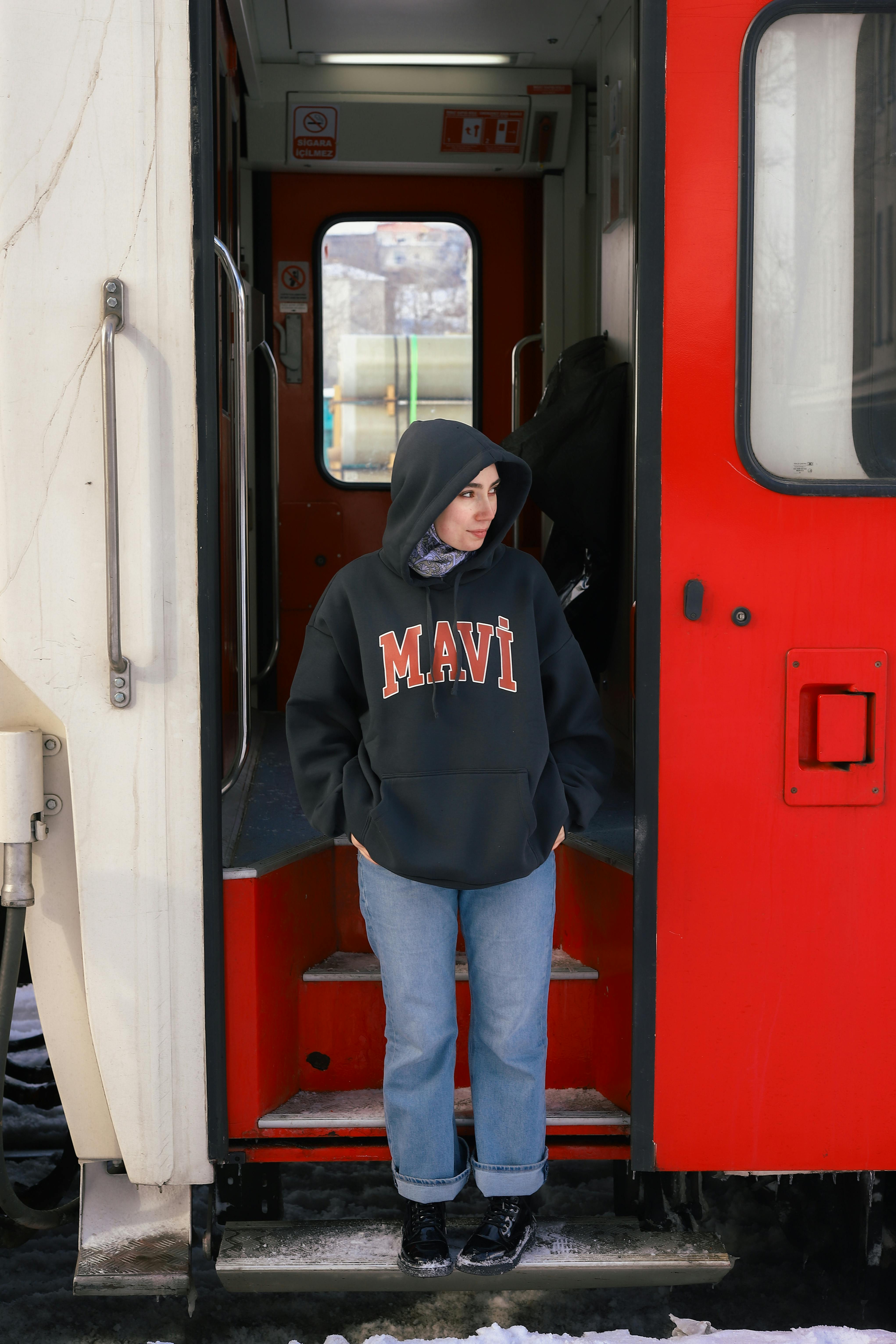 Person in a hoodie stands in train doorway with a snowy backdrop, looking contemplative.
