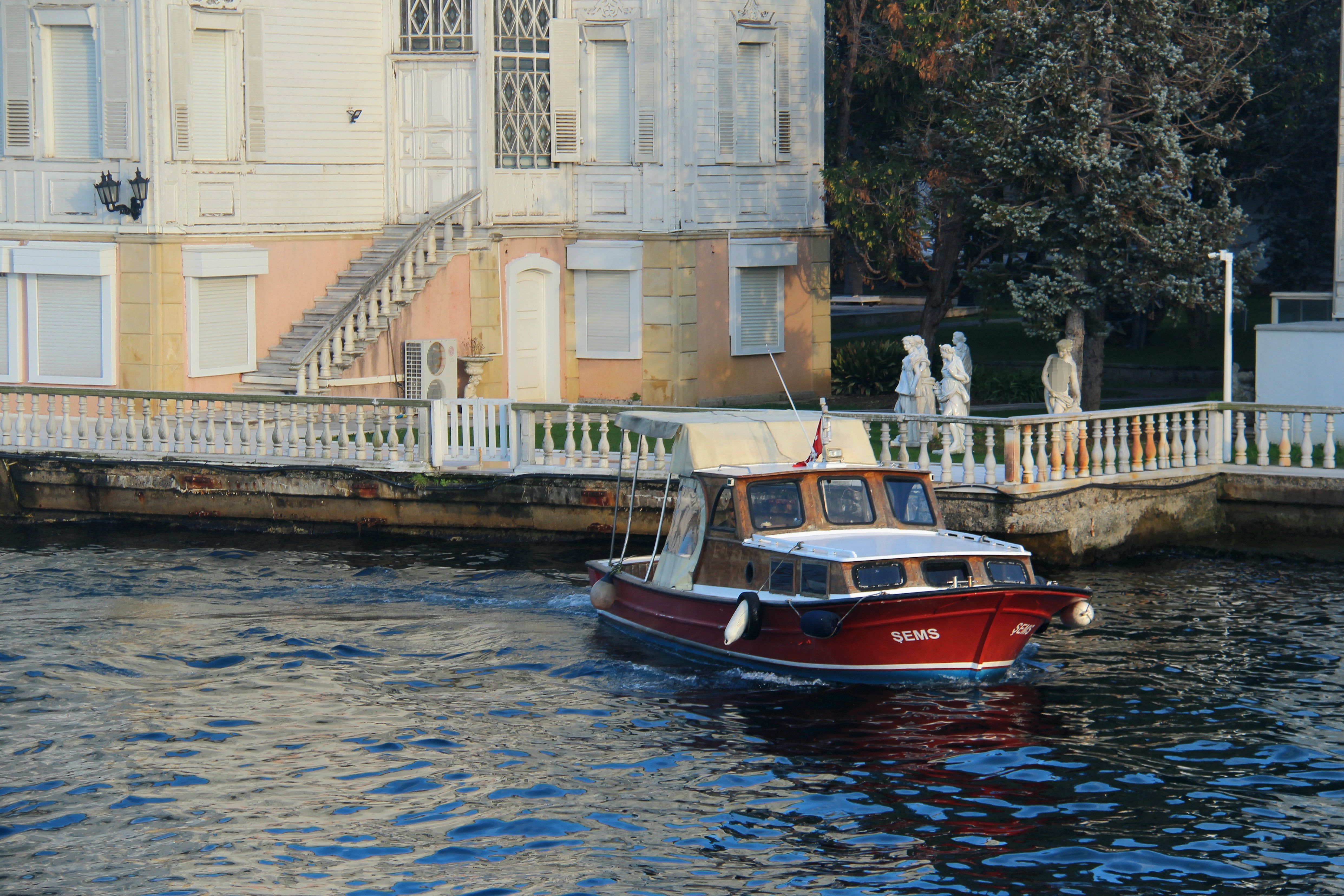A red boat cruising past a historic waterfront house on a sunny day.