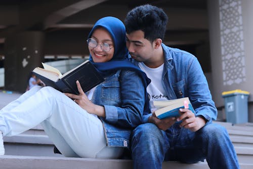 Man in Blue Denim Jacket Sitting Beside Woman in Blue Scarf Reading Book