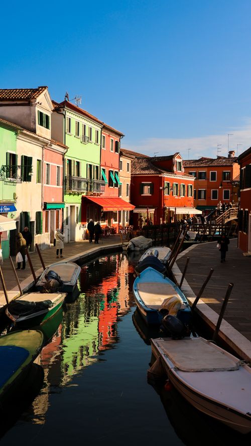 Free Charming view of vibrant houses lining a canal in Burano, Venice with boats and reflections. Stock Photo