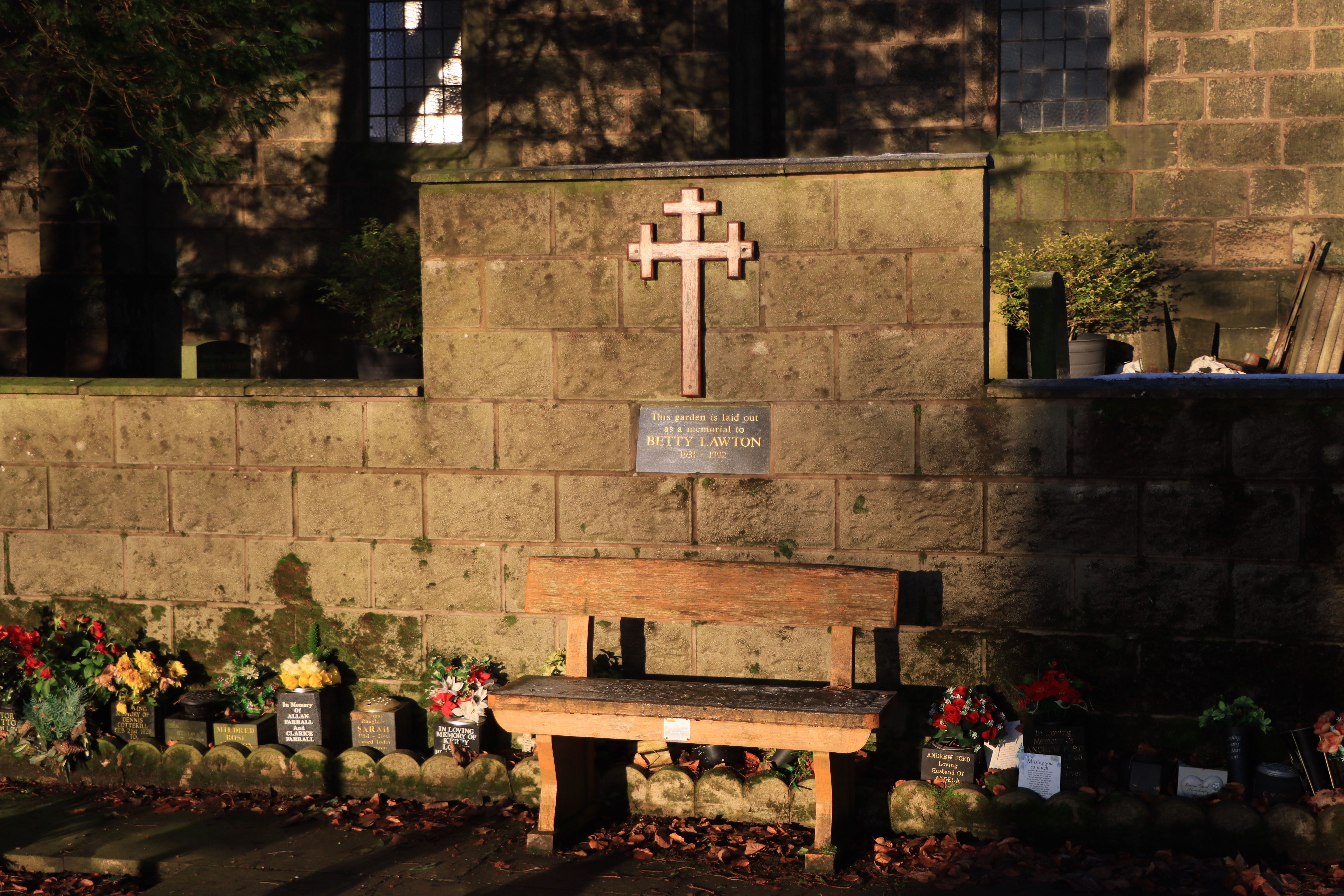 Tranquil cemetery scene with wooden bench, flowers and memorial plaque in sunlight.