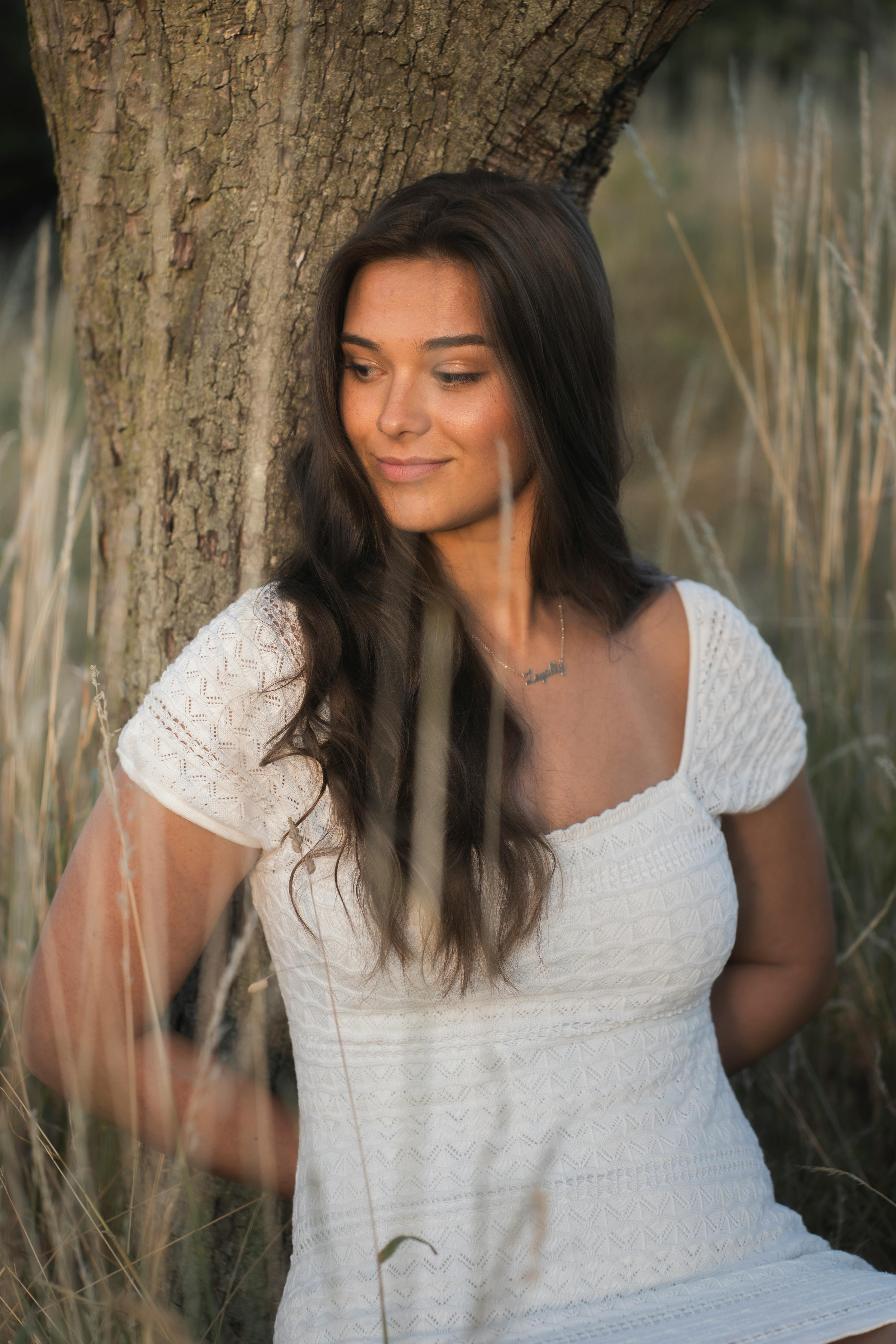 Bezpłatne Kobieta w białej sukni siedzi pod drzewem, emanując spokojem i naturalnym pięknem. Zdjęcie z galerii
