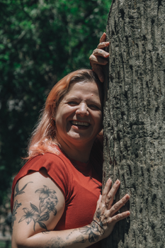 Joyful woman with tattoos hugging a tree in a sunny park setting.