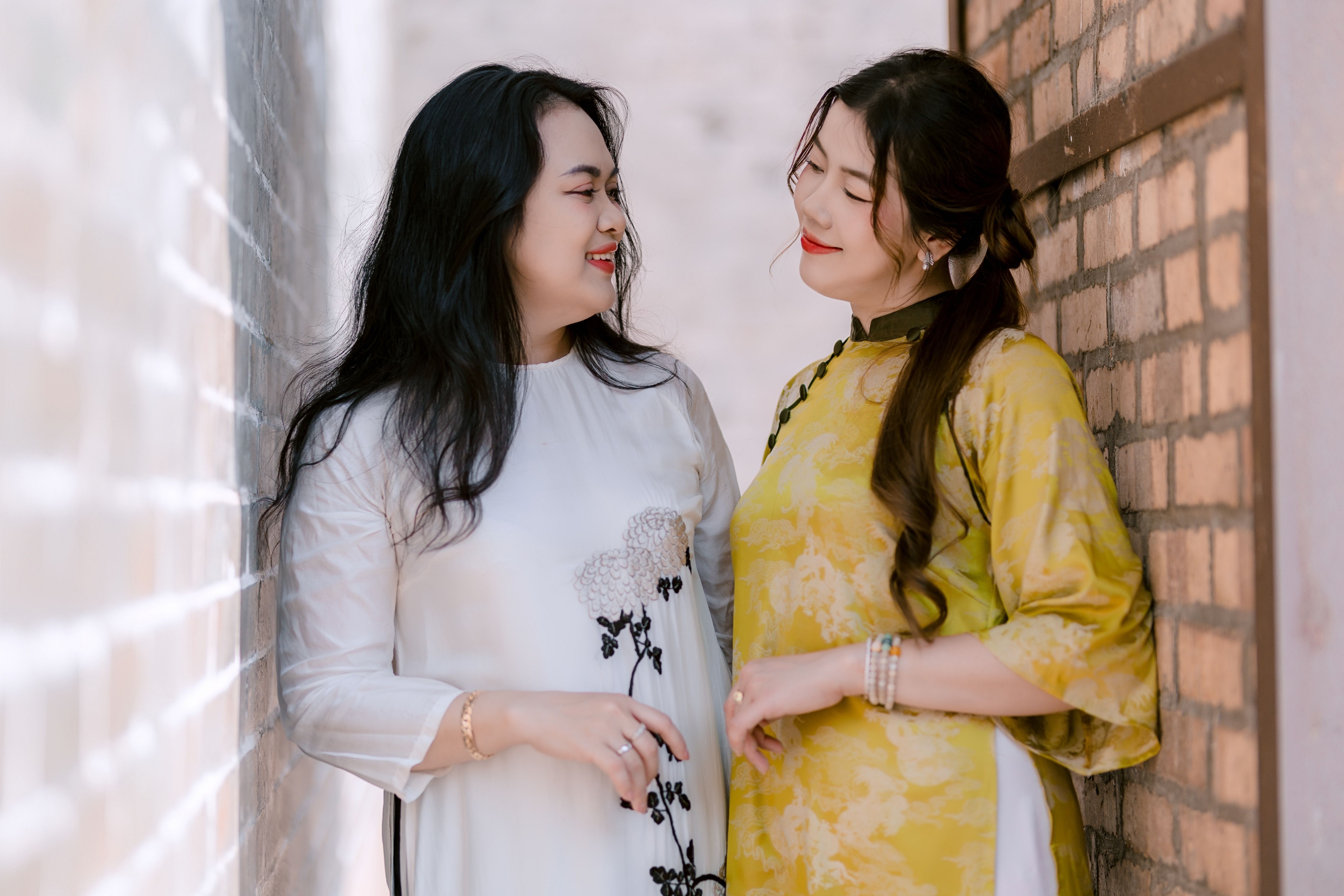 Two women smiling at each other in elegant traditional attire in a narrow brick alleyway.