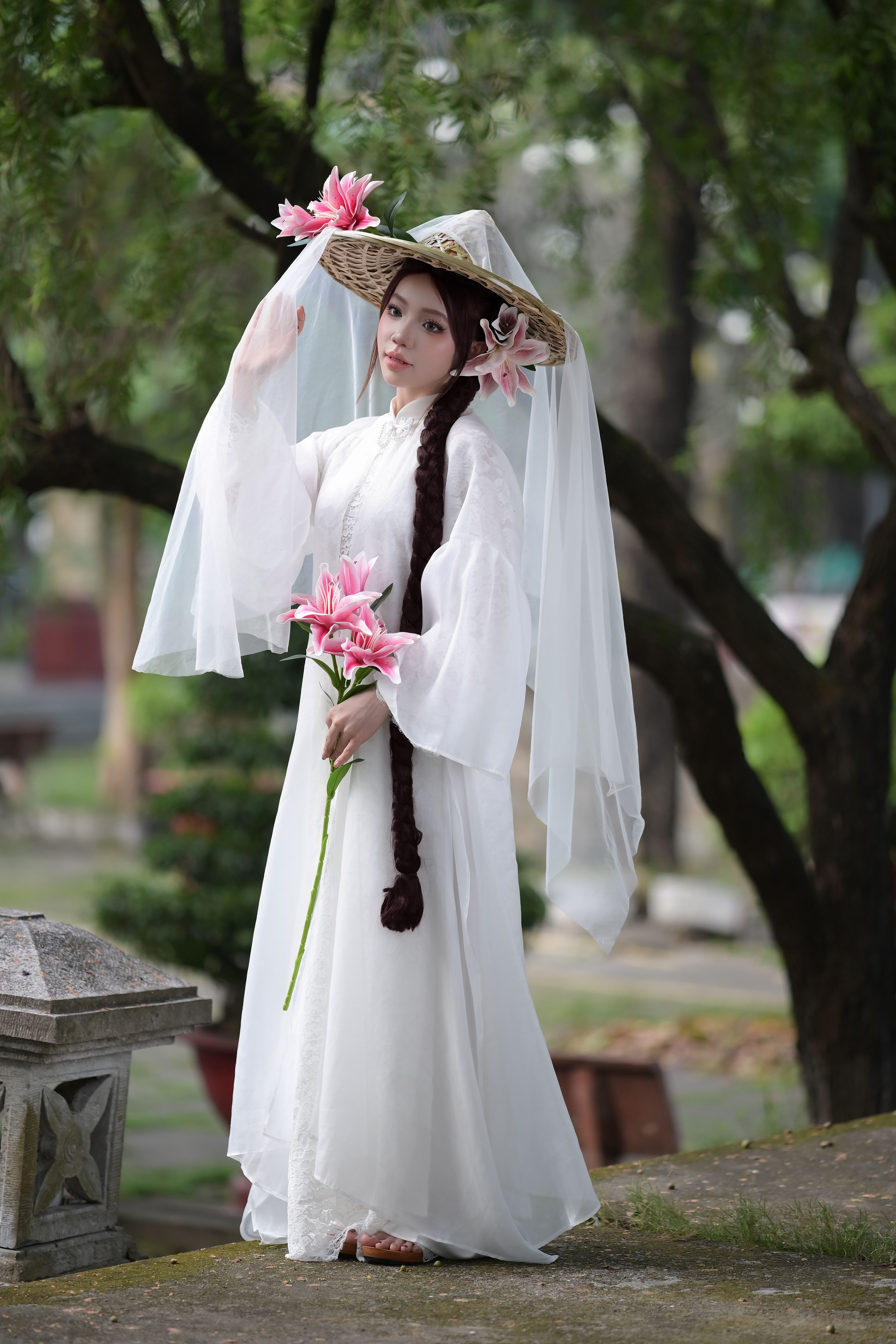 A woman in traditional attire with lilies and a woven hat stands outdoors capturing serenity.