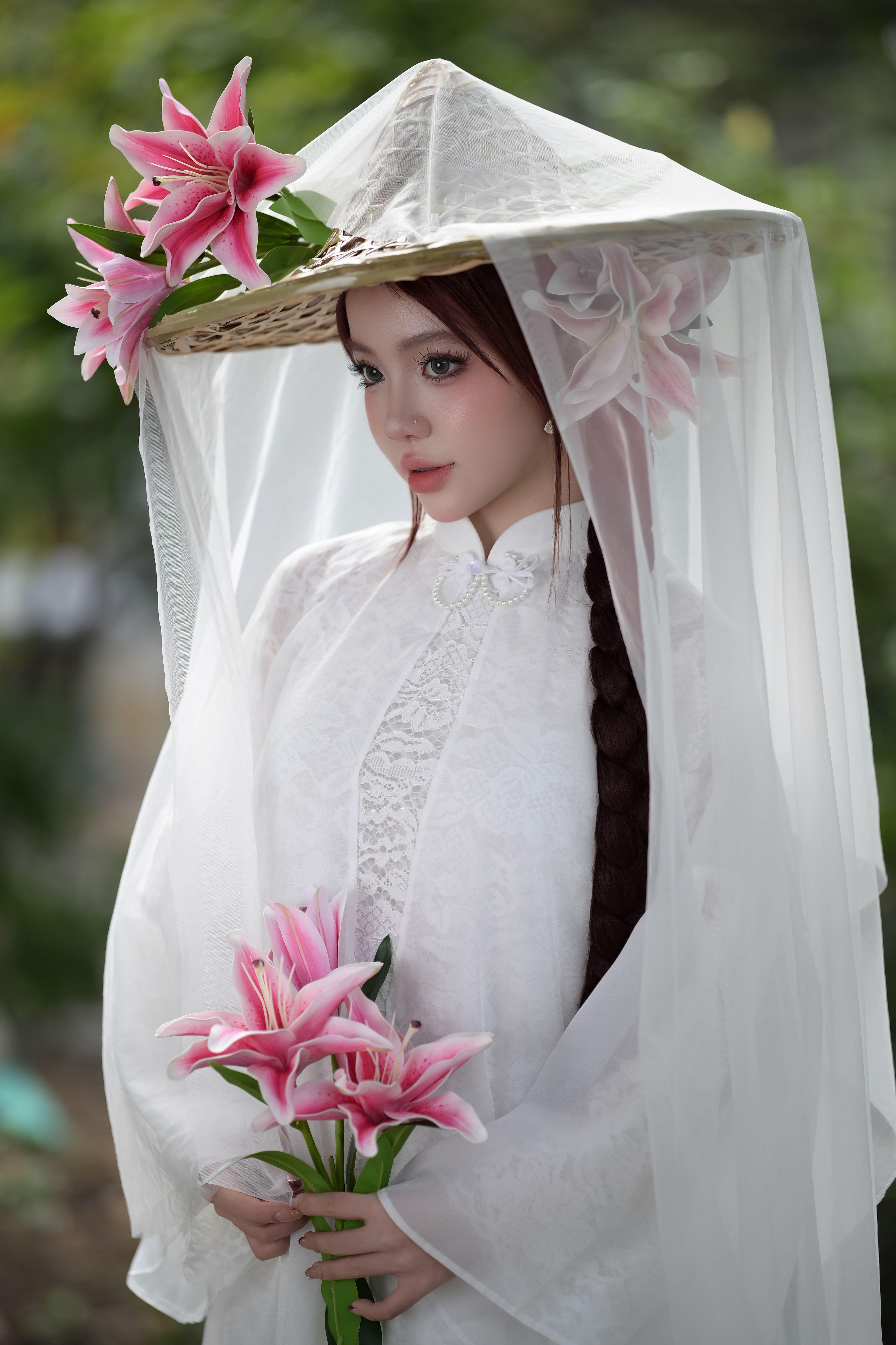 Capture of a woman in white traditional clothing holding pink lilies under a decorative hat.