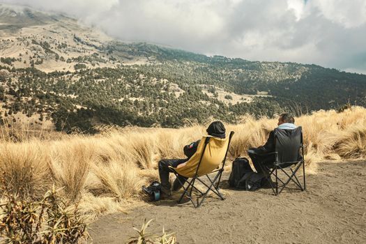 Two people relaxing in chairs amidst a vast, scenic mountain landscape under cloudy skies.