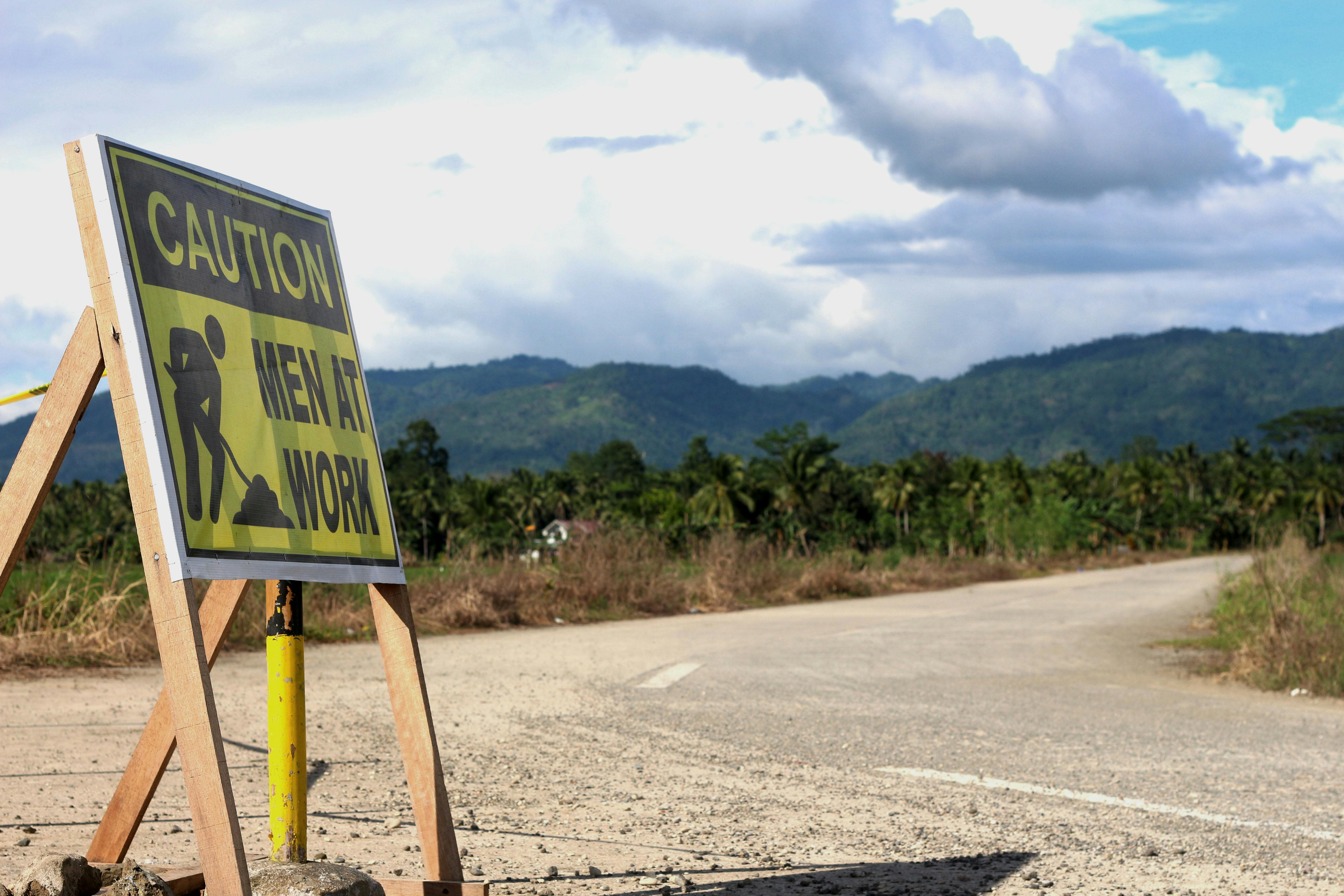A men at work sign on a rural road with a mountainous landscape in the background.