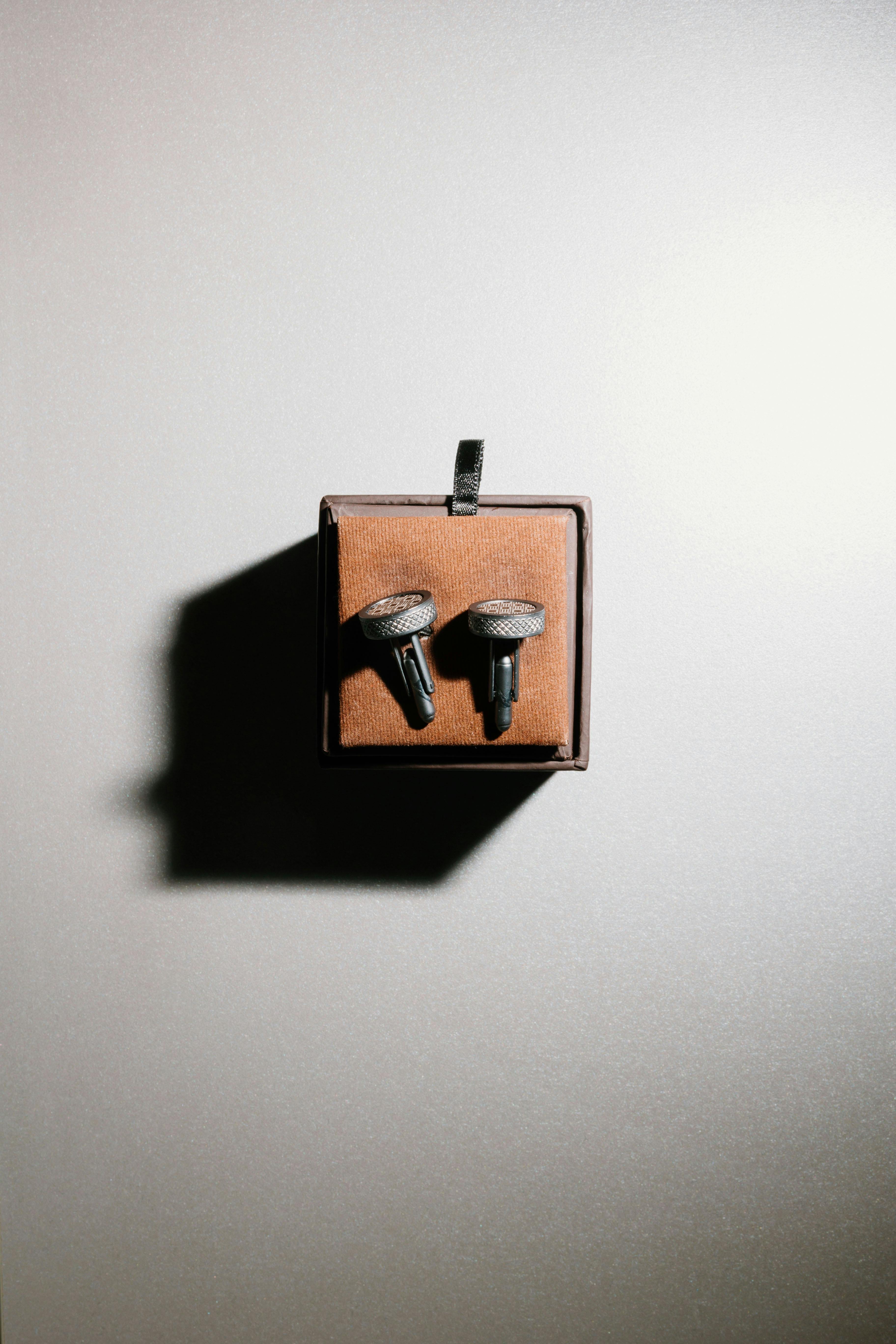 Free Minimalist presentation of black stud earrings in a brown box on white background, casting a distinct shadow. Stock Photo