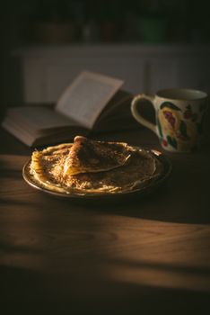 A plate of crepes in warm light, accompanied by a book and floral mug on a wooden table.