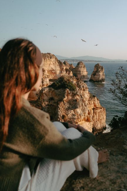 A woman sitting on a cliff admiring the rocky coastline and ocean during sunset.