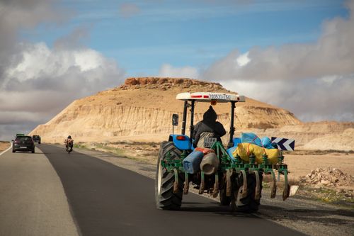 Free A tractor travels on a rural road in Morocco, passing by scenic desert landscapes under a blue sky. Stock Photo