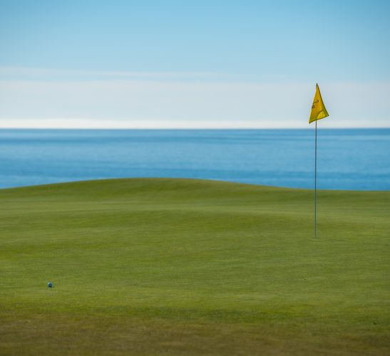 A coastal European golf green overlooking the sea, with a flag pin in the foreground and the Atlantic horizon behind it on a clear day.