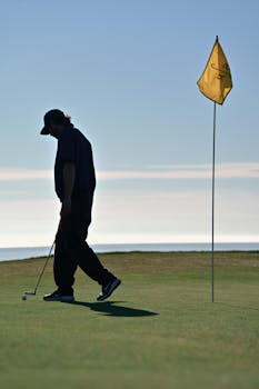 A golfer walking near a flag on a coastal golf course in the sunlight.