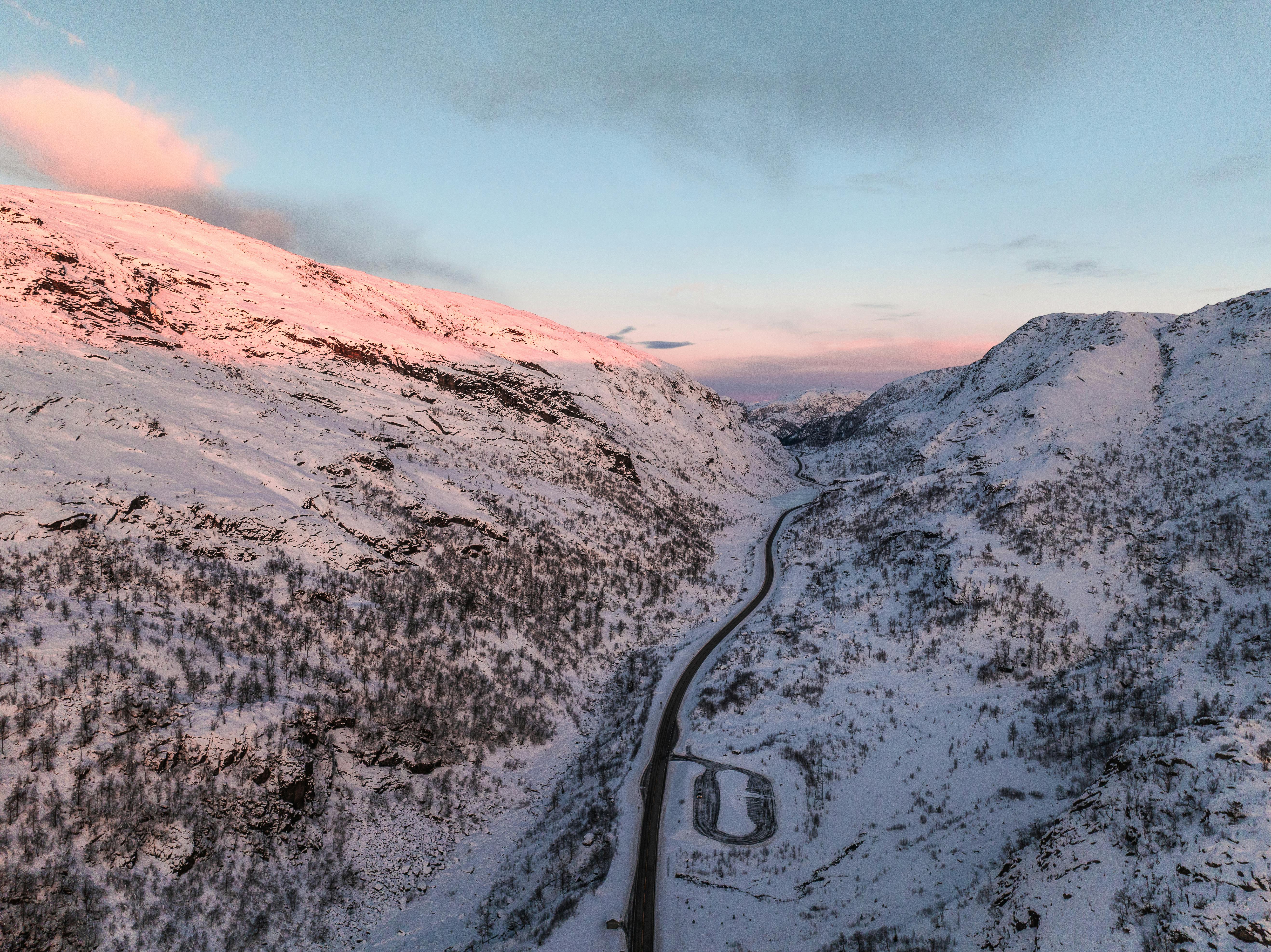 Aerial shot of snow-covered mountains and winding road in Norway during a stunning sunset.
