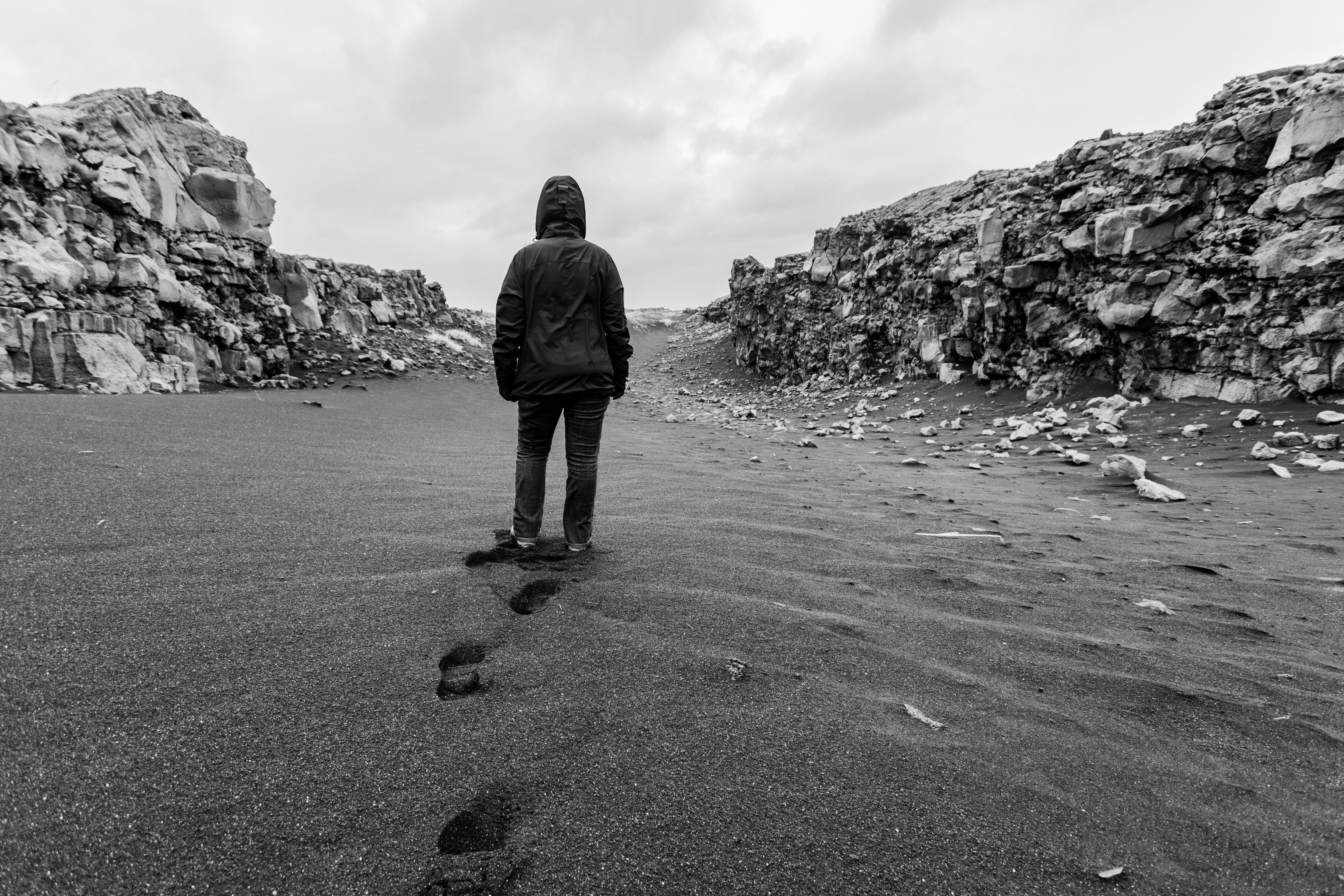 A person in a hooded jacket walks through a desolate rocky landscape, leaving footprints in the sand.