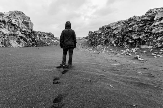 A person in a hooded jacket walks through a desolate rocky landscape, leaving footprints in the sand.