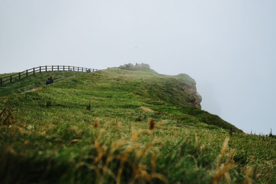 A serene and misty cliffside landscape with lush green grass and a distant view of hikers.