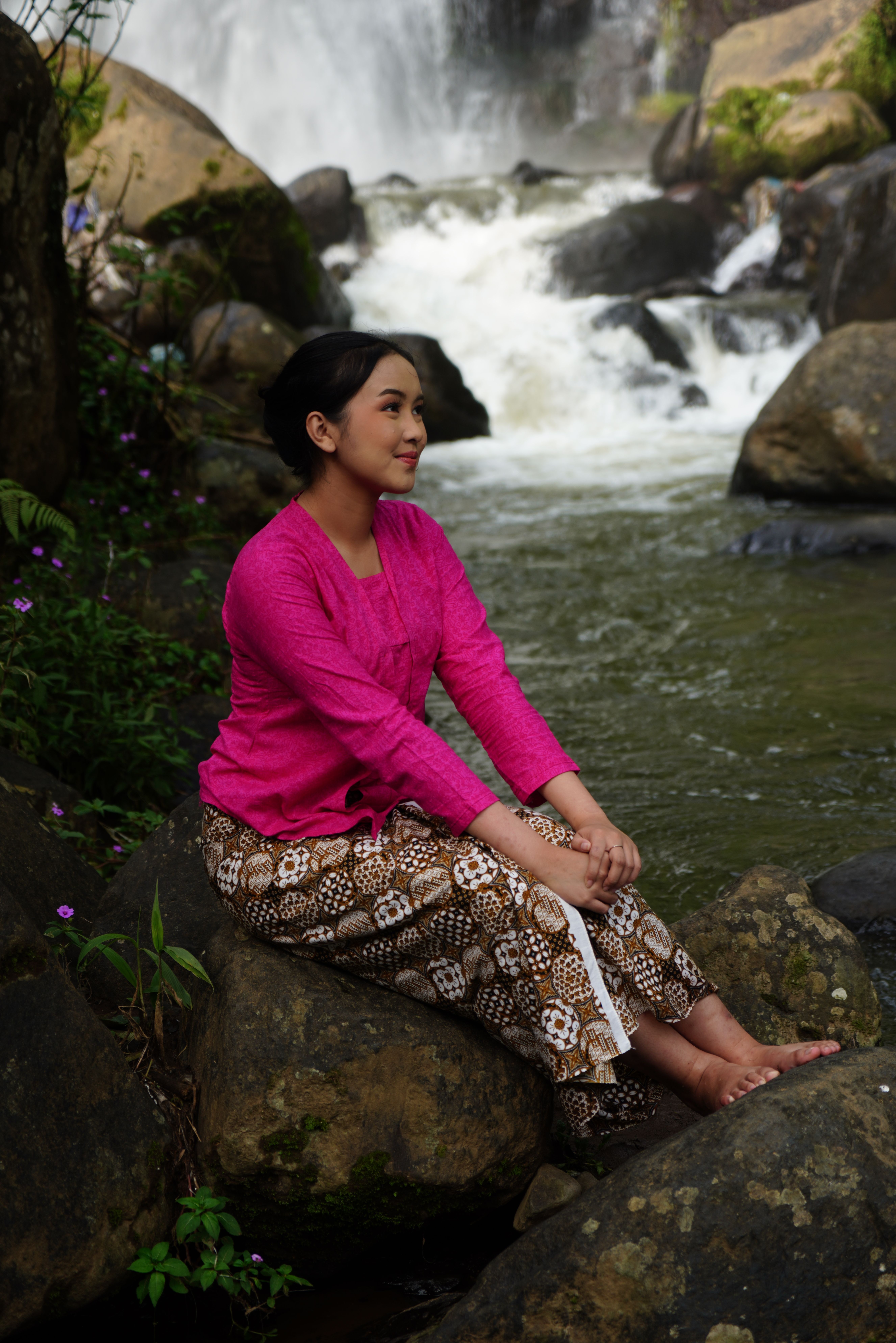 A serene moment captured of a woman in traditional attire seated by a waterfall in West Java.