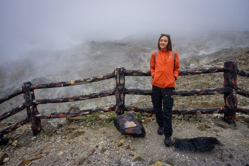 Free Woman standing on Mount Papandayan, Indonesia, in foggy weather, wearing an orange jacket and enjoying the outdoor scenery. Stock Photo