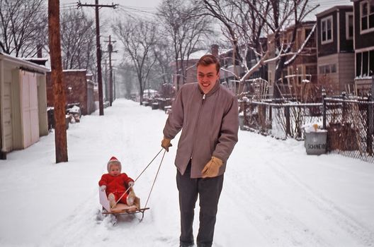 Father in winter attire pulling child on sled through snowy neighborhood alley.