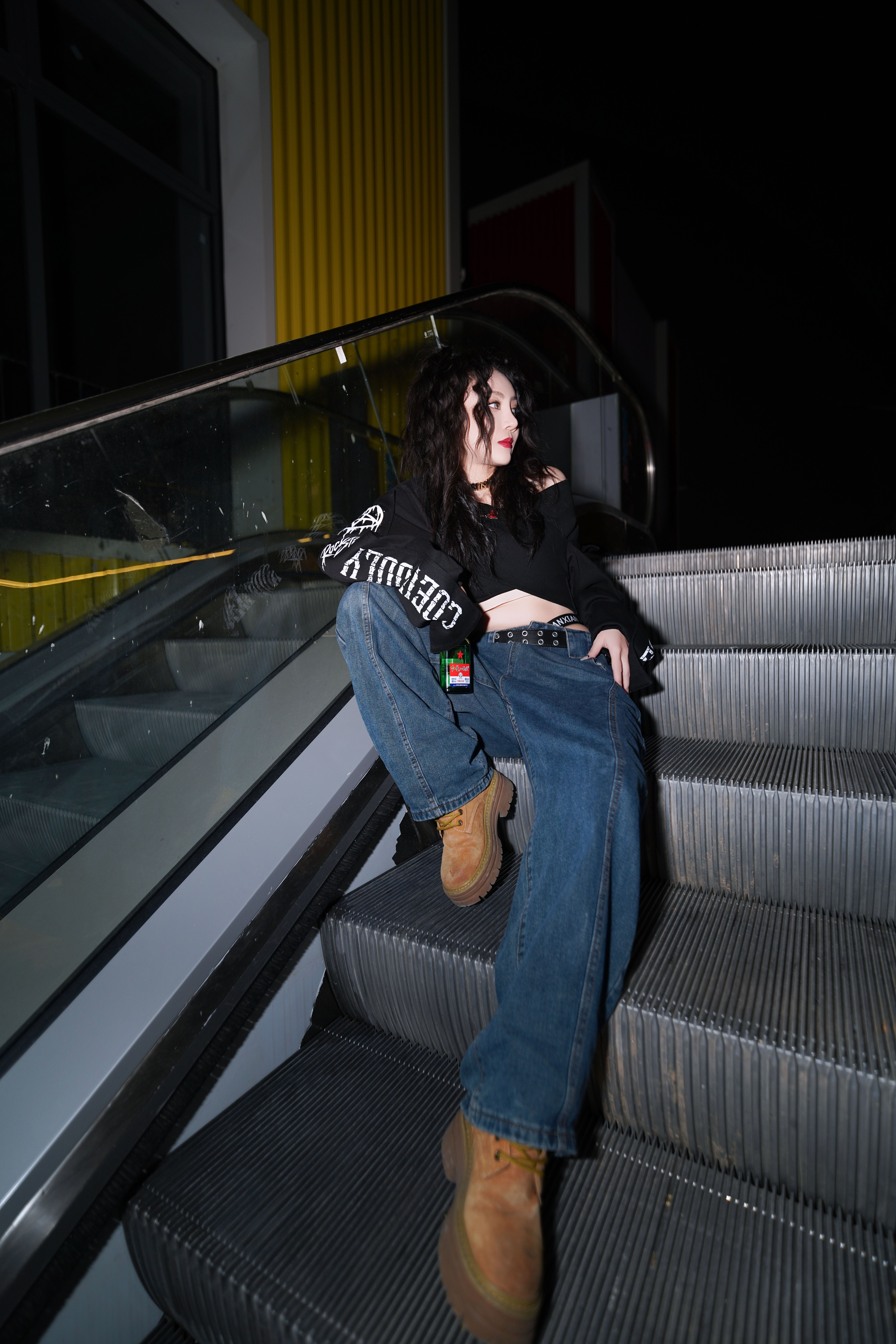 Stylish young woman poses on escalator steps with bold streetwear fashion at night.