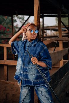 Young adult in denim with sunglasses poses at rustic outdoor setting.