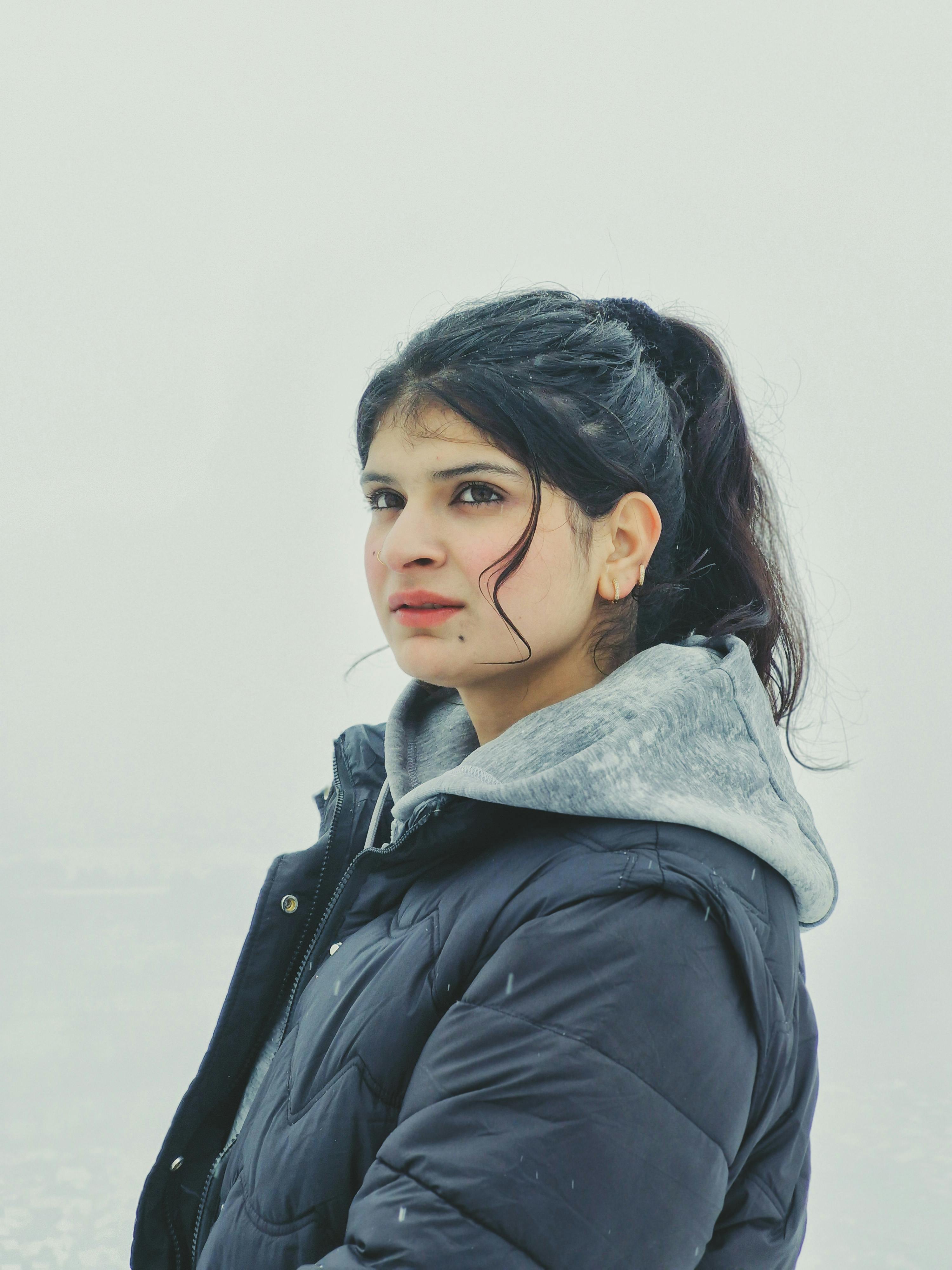 A young woman in a winter coat stands outdoors in a snowy landscape, gazing thoughtfully.