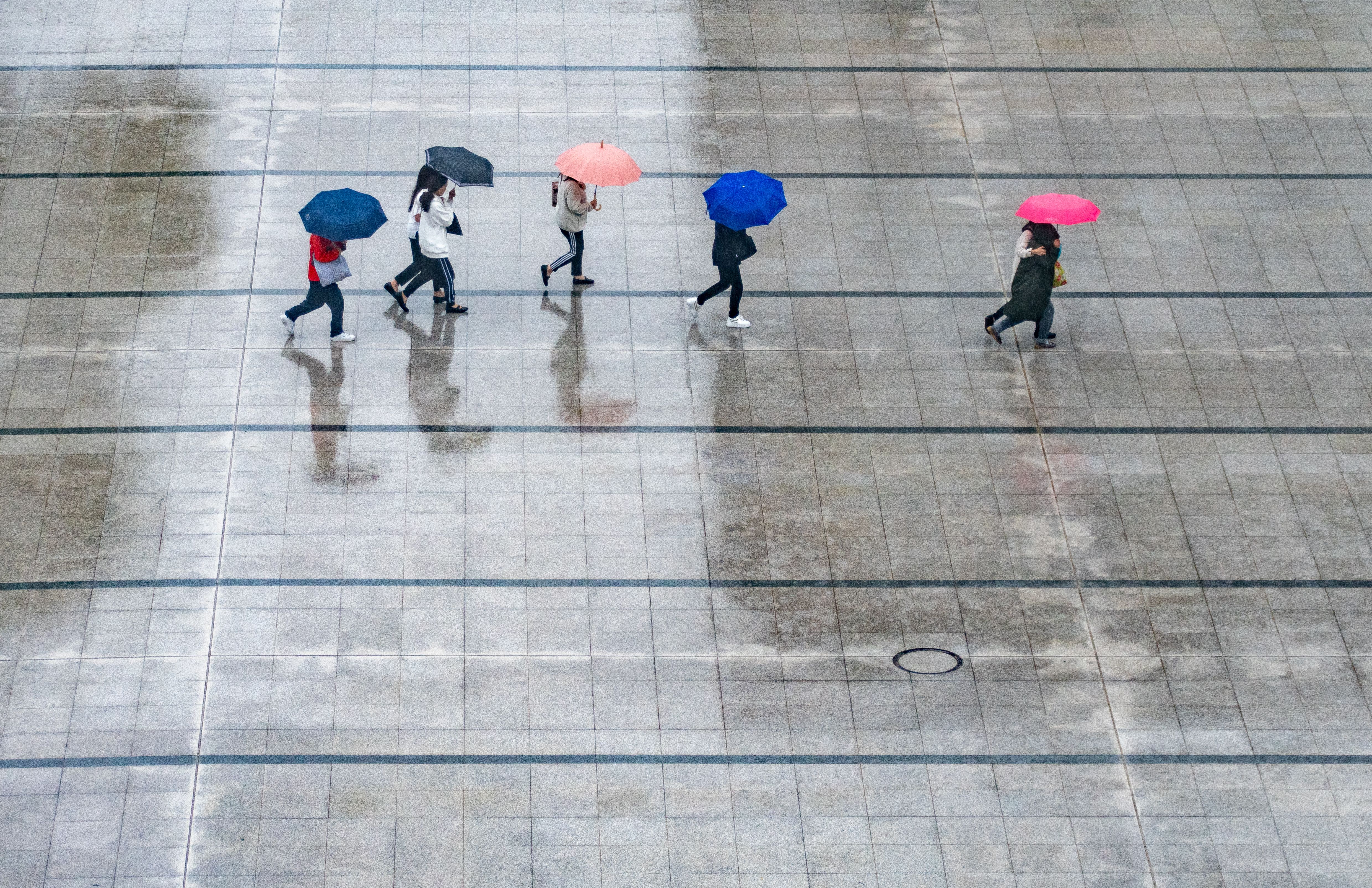 Aerial view of people walking with umbrellas on a rainy day, reflecting in wet pavement.