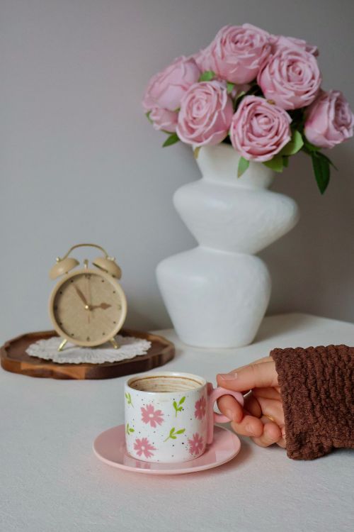 Free A hand holds a floral coffee cup beside a vase of pink roses and a vintage clock. Stock Photo