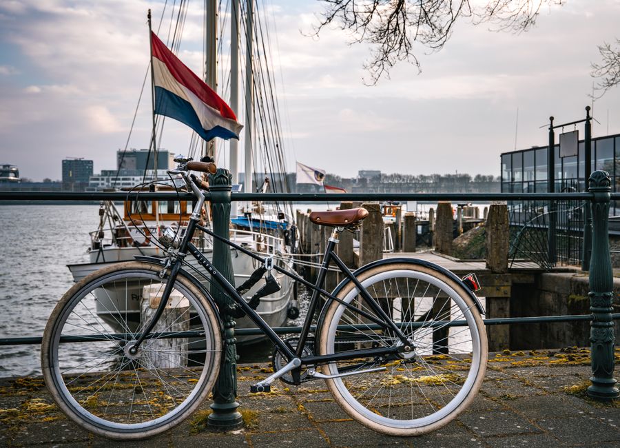 A bicycle parked by a harbor in the Netherlands with boats and the Dutch flag visible.