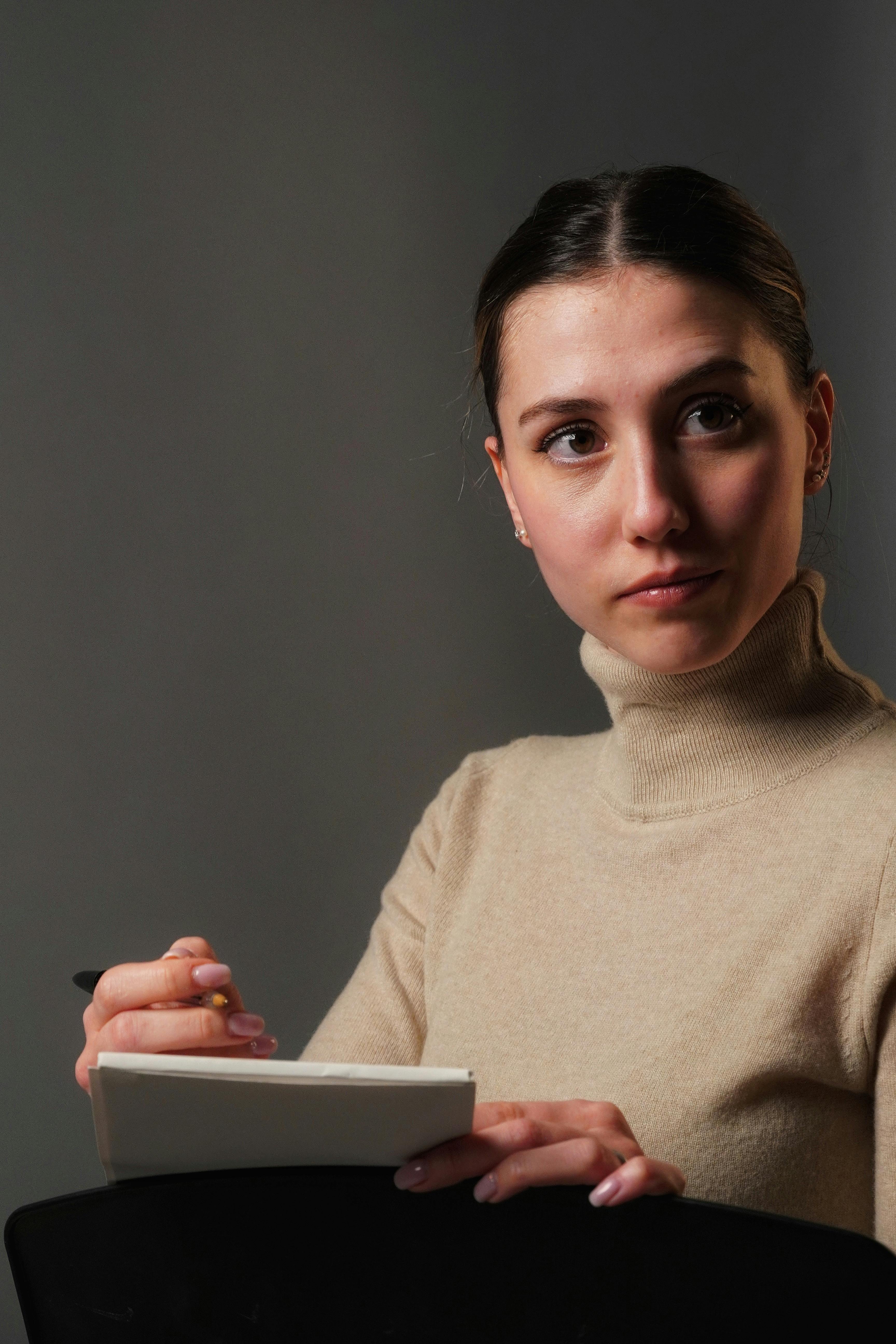 Gratis Una donna con un dolcevita beige tiene in mano un quaderno e una penna e guarda pensierosa di lato. Foto a disposizione