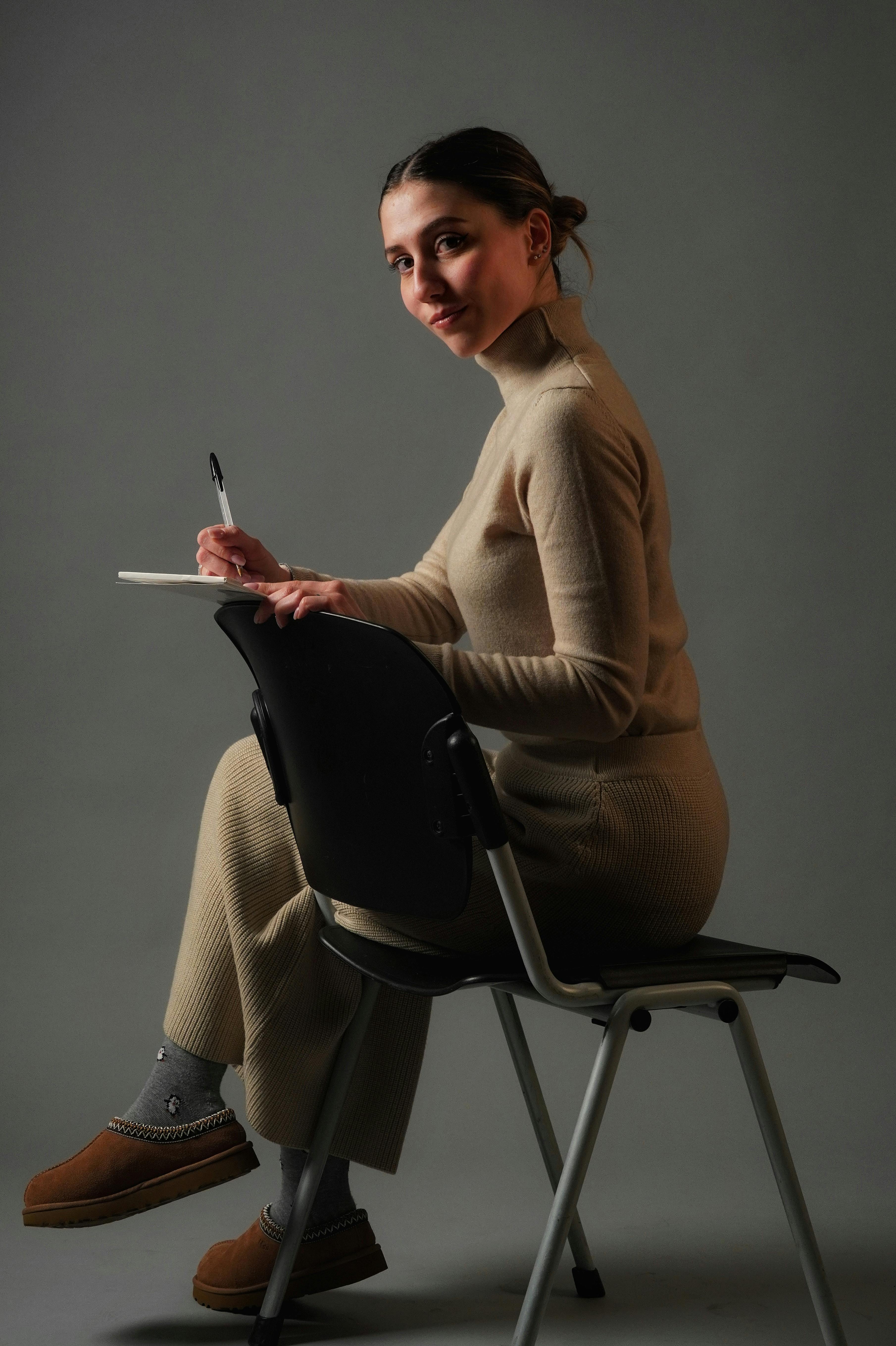 Gratis Una donna vestita di beige scrive pensierosa in uno studio, seduta comodamente su una sedia. Foto a disposizione