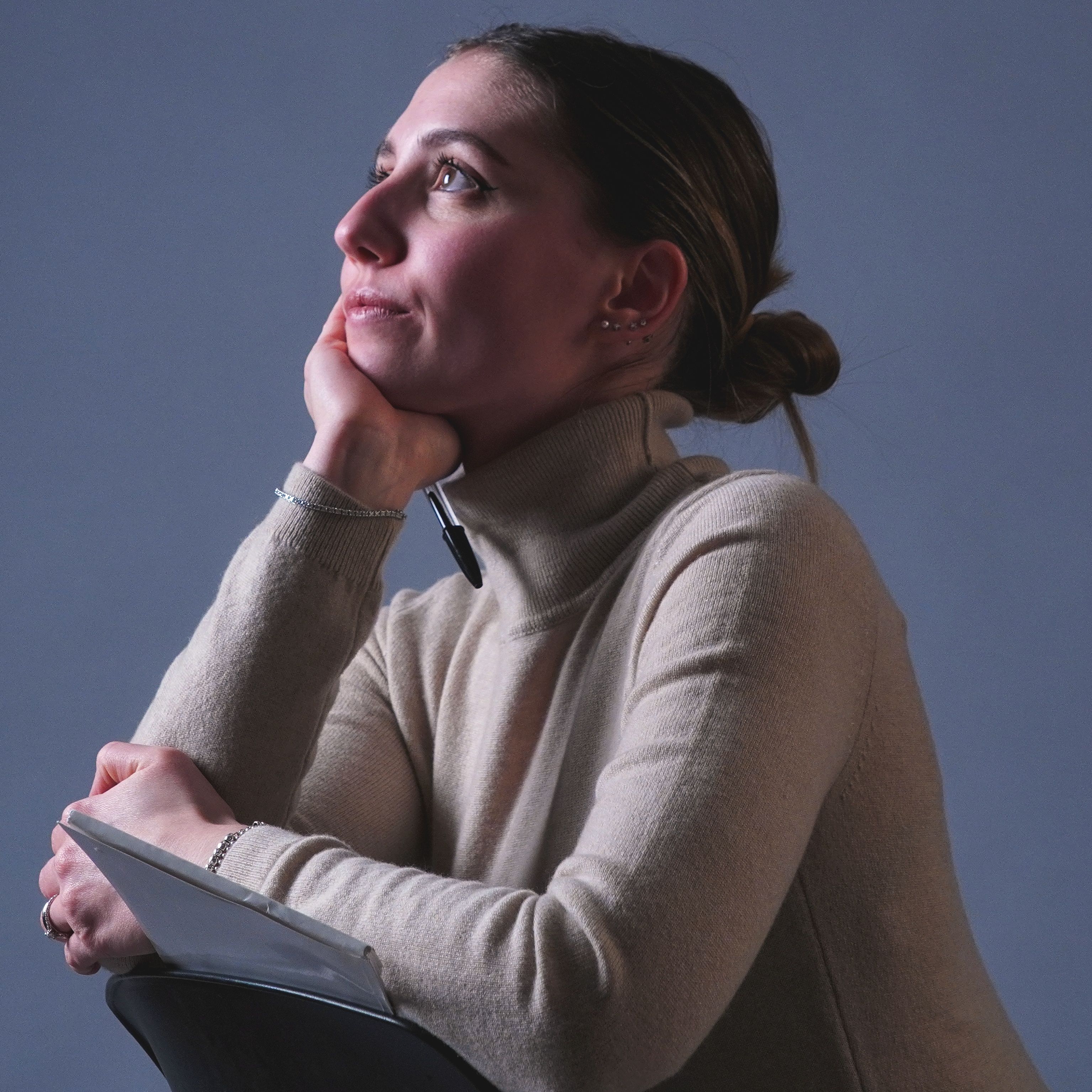 A thoughtful woman in a cozy sweater gazes upwards, holding a book, indoors.
