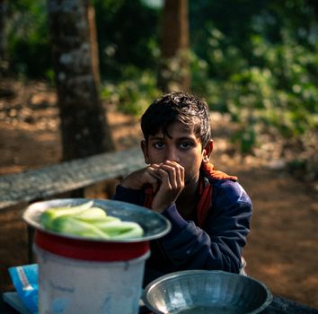 A young boy sits at a table outdoors with food items, deep in thought.