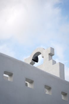 A minimalist white bell tower against a cloudy blue sky, showcasing Greek architecture.