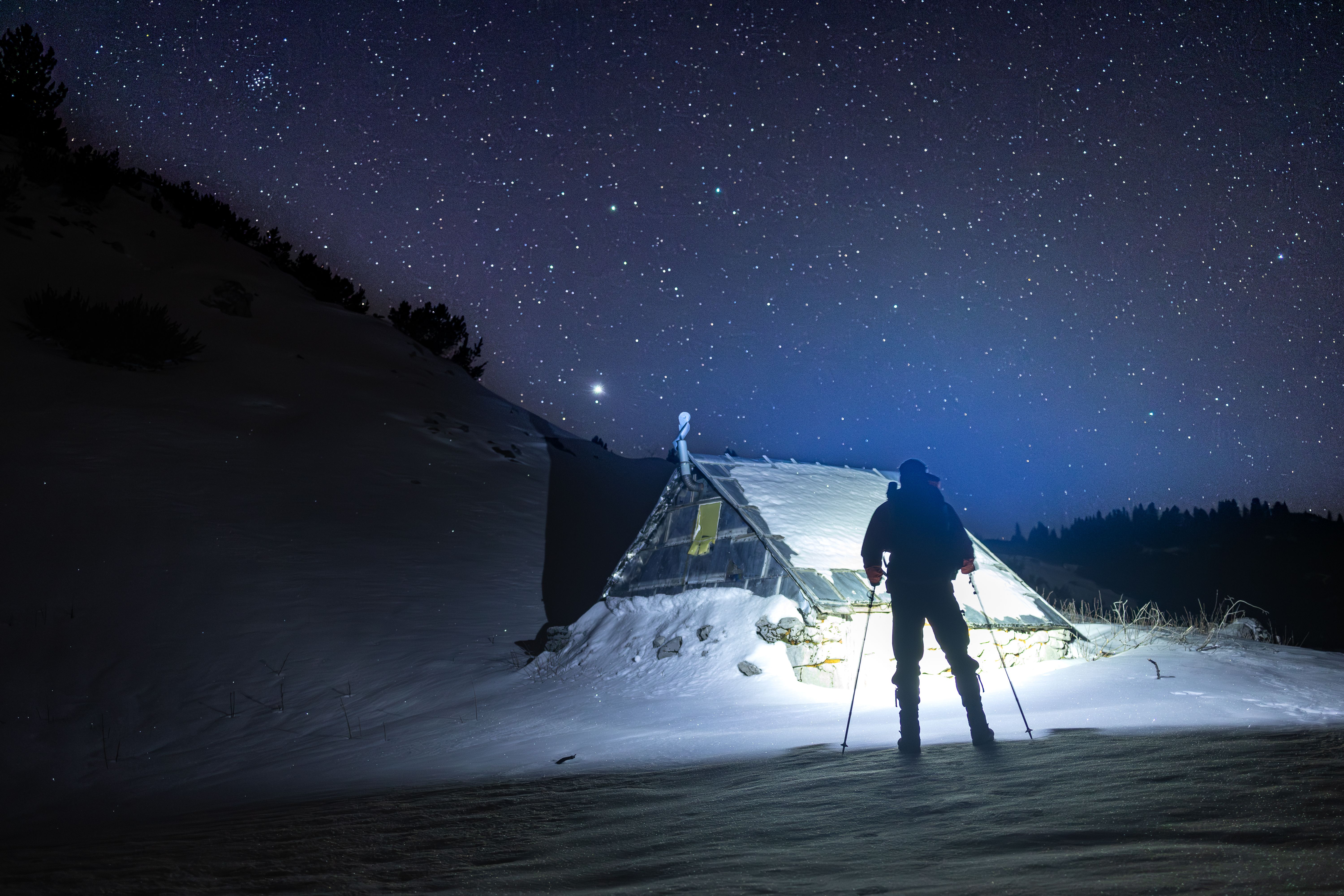 Silhouette of a skier standing by a snow-covered tent under a starry night sky in winter.