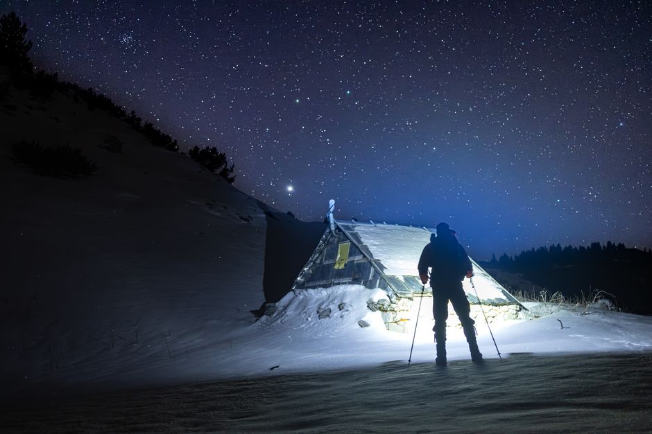Silhouette of a skier standing by a snow-covered tent under a starry night sky in winter.