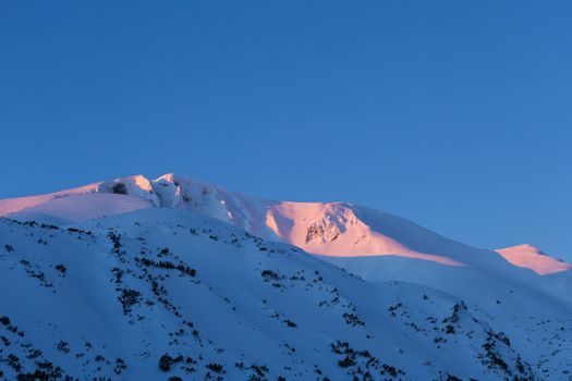 Stunning view of snow-covered mountains with pink sunrise glow against a clear blue sky.