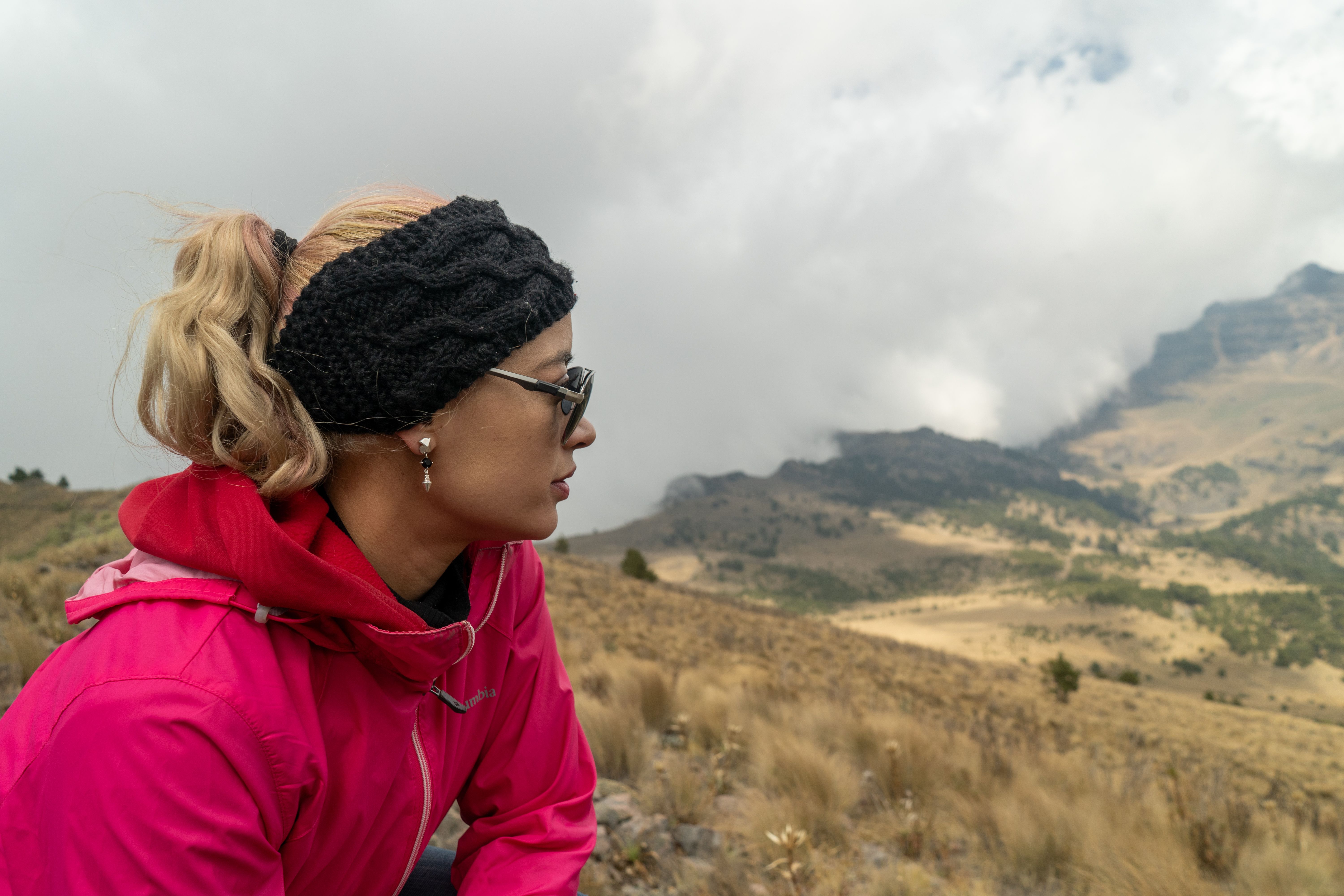Woman wearing pink jacket hiking in mountainous terrain with cloudy sky.