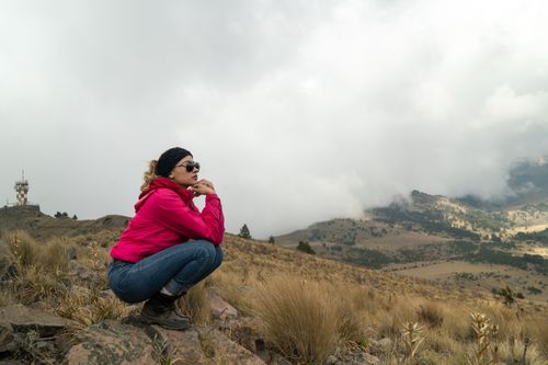Free A woman in a pink jacket crouches on a rocky mountain with a cloudy view. Stock Photo
