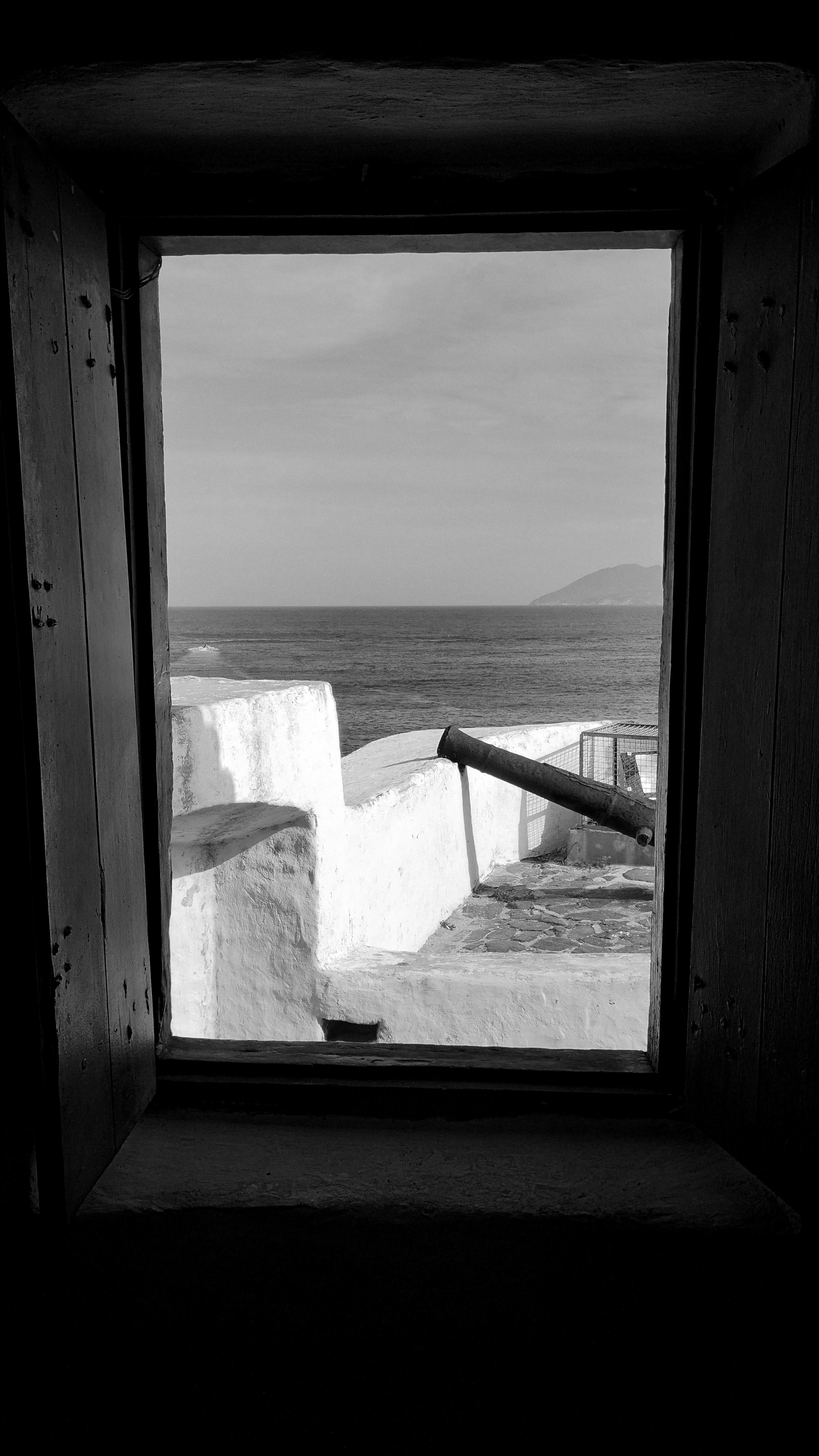 Monochrome view through a window featuring a historic cannon overlooking the Cabo Frio coastline.