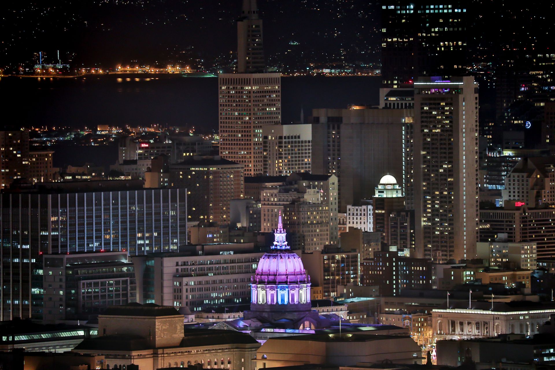 San Francisco downtown skyline at night featuring the illuminated City Hall dome, surrounding skyscrapers, and bay waterfront
