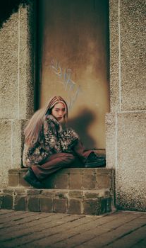 Artistic portrait of a young woman in fur coat sitting on steps in urban setting.