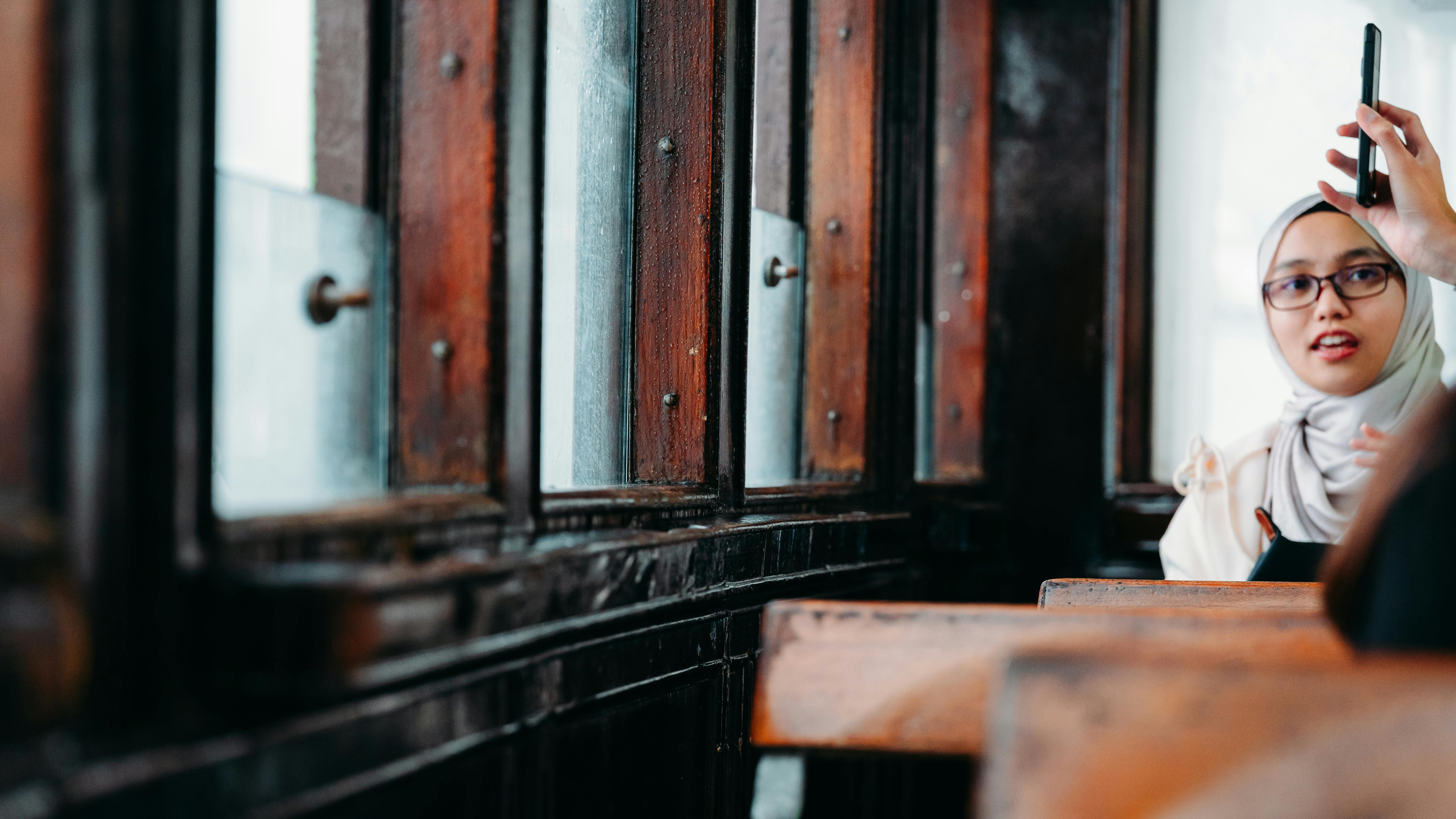 grátis Uma mulher de hijab tirando uma selfie em um café aconchegante, com reflexos em painéis de madeira. Foto profissional